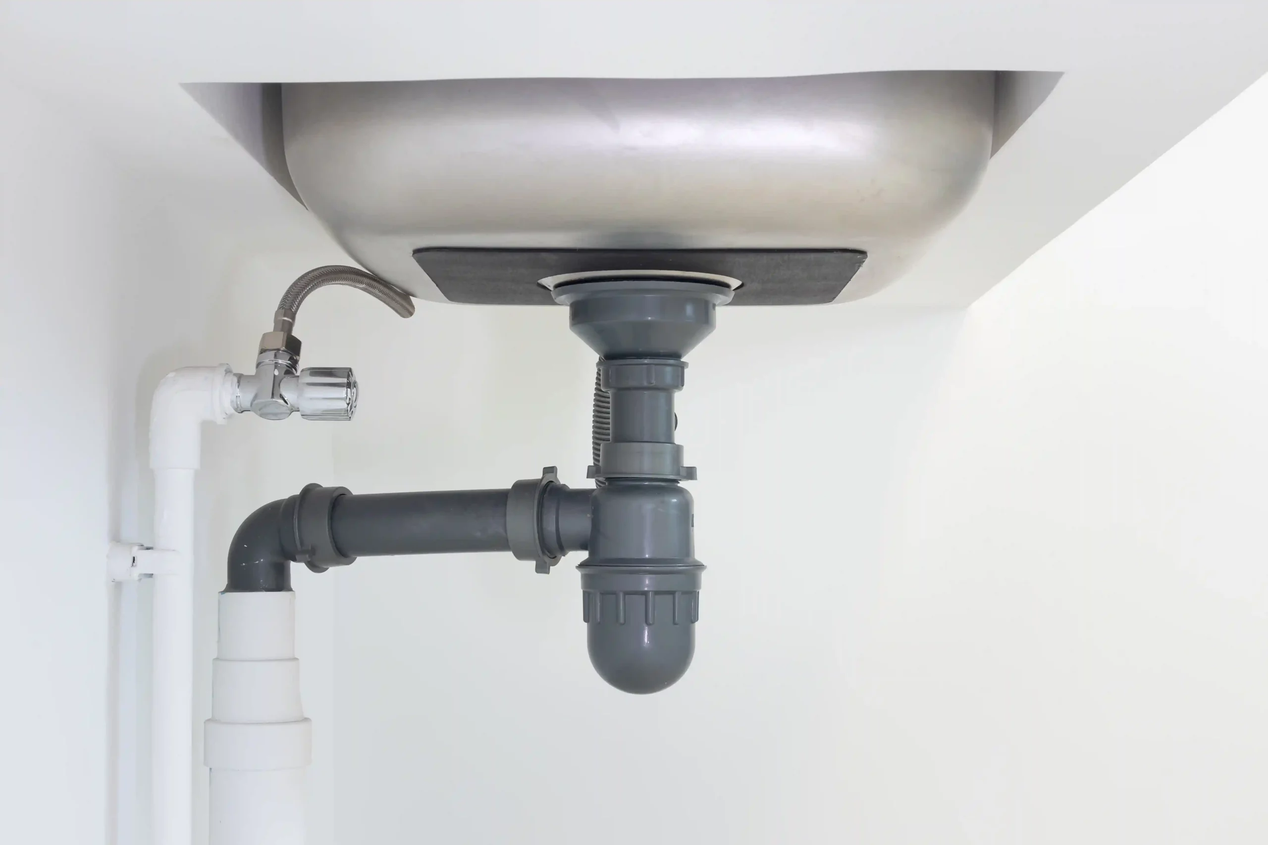 Grauer Flaschensiphon aus Kunststoff unter einem Spülbecken aus Chromstahl, mit Abgangsrohr zur Wand und abschraubbarem Becher unten